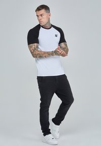 Camiseta de manga corta con cuerpo blanco y mangas raglán negras, con un pequeño logotipo bordado en el pecho, combinada con jeans negros y zapatos blancos.
