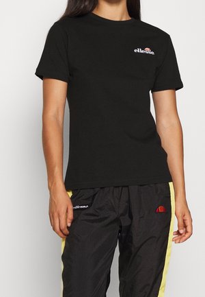 Kobieta ubrana w czarny t-shirt ellesse i czarne spodnie dresowe ellesse z żółtymi bocznymi paskami, stojąca na białym tle.