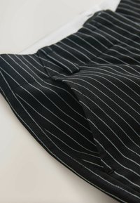 Čierne látkové nohavice obsahujú biele vertikálne pruhy, majú hladkú textúru a záhyb v páse. Viditeľný detail vrecka na boku.