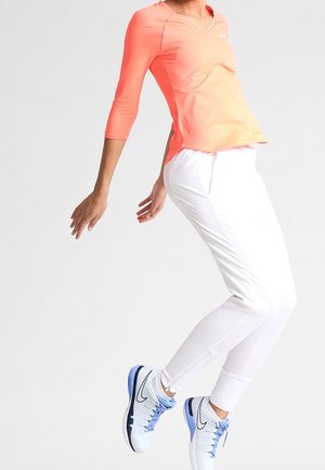 Persoon draagt een koraalroze top met lange mouwen, witte broek en witte-blauwe Nike sneakers, vastgelegd halverwege een sprong tegen een effen lichte achtergrond.