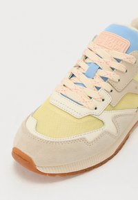 Baskets beiges et jaunes avec des textures en mesh et en daim, présentant des accents bleu clair, des lacets roses et une étiquette de marque rose sur la languette.