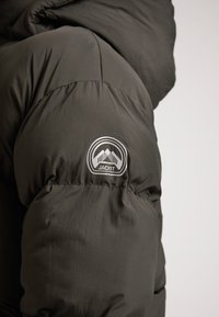 Giacca puffer verde scuro con un grande logo rotondo etichettato "JACKIT" e grafiche montane. Il tessuto sembra liscio e leggermente lucido.