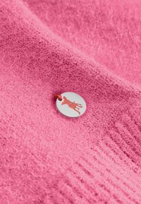 Różowy materiał o teksturze z okrągłą, srebrną nalepką przedstawiającą sylwetkę czerwonego psa, przymocowaną czerwoną nitką.