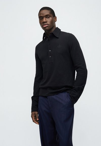 Άνδρας φορώντας μαύρο πόλο με μακριά μανίκια και μπλε πουά παντελόνι, στέκεται μπροστά σε απλό ανοιχτό γκρι φόντο.