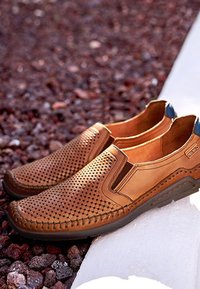 Braune Leder-Slipper mit perforiertem Design und schwarzen Sohlen, auf einem weißen Sims über rötlichem Kiesboden platziert.