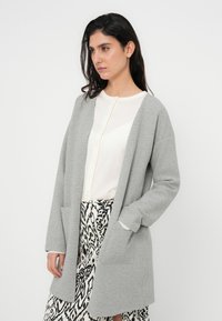 Γκρι ζακέτα με άνετη γραμμή, μακριά μανίκια, ανοιχτό μπροστινό μέρος και δύο πλαϊνές τσέπες, φορεμένη πάνω από ένα ανοιχτό κρεμ τοπ και φούστα με ασπρόμαυρο μοτίβο.