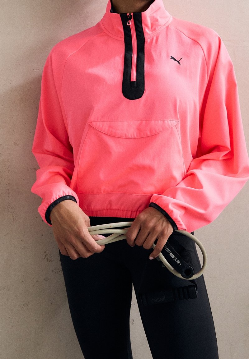 Vaaleanpunainen neuletakki, jossa on korkea kaulus, musta vetoketju ja etutasku. Mustat yksityiskohdat hihansuissa, yhdistettynä mustiin legginsseihin ja kuntoiluvälineisiin.