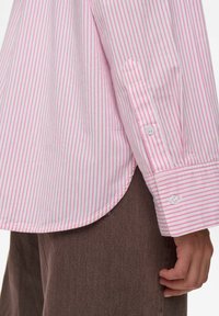Gestreept shirt met roze en witte verticale strepen, lange mouwen, enkele knopen bij de manchetten en een gebogen zoom. Geddragen met bruine textuurbroek.