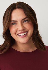 Close-up of a person with shoulder-length, wavy brown hair, wearing a burgundy top. Skin has a natural tone and a slight sheen.