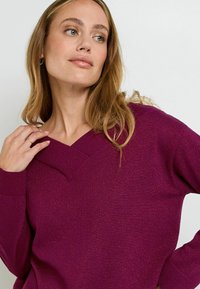 Maglione a V in magenta scuro con polsini e orlo a coste. Tessuto morbido con una leggera lucentezza. Modella posa con una mano sulla spalla.