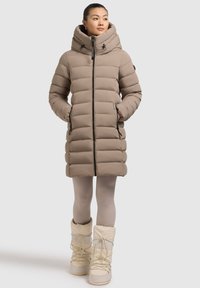 Beige Daunenjacke mit großer Kapuze, Reißverschluss und vertikalen Nähten. Getragen über leichten Leggings und weißen, schnürbaren Winterstiefeln.