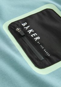 Šviesiai mėlynas audinys su kontrastinga juoda kišene, kuri turi užtrauktuką ir baltą tekstą "BAKER by TED BAKER". Apvalūs kišenės kraštai.