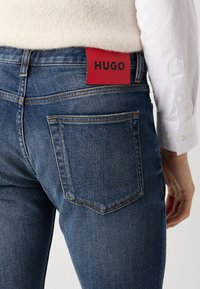 Tamno plave traperice s crvenom HUGO etiketom na struku, s zlatnim šavovima i standardnim dizajnom stražnjih džepova.
