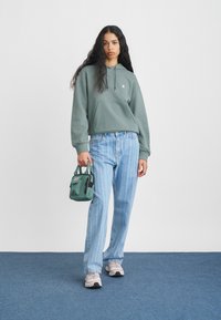 Groene hoodie, lichtblauwe gestreepte jeans en grijze sneakers. Houdt een kleine groene tas vast. Rechte snit, ontspannen pasvorm en zachte textuur.