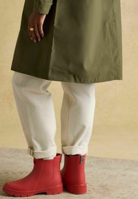 Bocanci roșii din cauciuc pentru gleznă cu tălpi texturate, asortați cu pantaloni crem suflecați și o haină verde măsliniu. Accent pe un design practic, rezistent la intemperii.