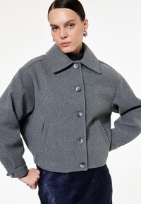 Grå uldblandet cropped jakke med stor krave, knaplukning og to sidelommer. Accentueret af runde grå knapper. Tekstureret stof.