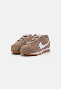 Smeđe antilop tenisice s bijelim Nike swooshom, okrugli vrh, ravne vezice i gumeni potplat. Mekana tekstura, retro dizajn.