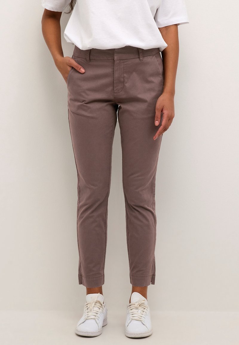 Femme portant un pantalon marron ajusté, un t-shirt blanc et des baskets blanches, debout avec une main dans la poche devant un fond uni.
