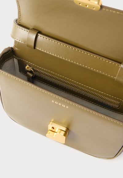 Χειροποίητη τσάντα ώμου από δέρμα σε ελιά χρώμα με χρυσό κούμπωμα, ορατή τσέπη με φερμουάρ στο εσωτερικό και το όνομα της μάρκας «soeur» εγχάρακτο στην μπροστινή όψη.