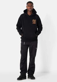 Sudadera negra con un bolsillo frontal, que presenta un diseño gráfico dorado en el pecho. Combinada con pantalones cargo negros y zapatillas negras.