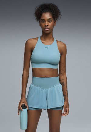 Lyseblå sportstop og matchende højtaljet lagdelt nederdel med shorts under. Tekstureret vandflaske holdt i højre hånd.