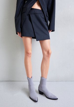 Kvinde iført en mørk asymmetrisk nederdel, matchende blazer og grå ankelstøvler med spids tå, stående mod en ensfarvet væg.