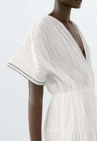 Femme portant une robe blanche plissée à manches courtes avec un bord noir sur les manches et un décolleté en V profond.