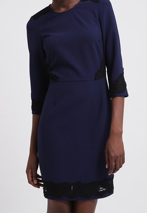Fitted navy dress with three-quarter sleeves and black lace trim on shoulders, cuffs, waist, and hem, worn by a standing person.