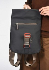 Mochila de tela negra con un bolsillo frontal con cremallera, una correa de acento naranja y correas de hombro marrones ajustables. Presenta una textura suave.