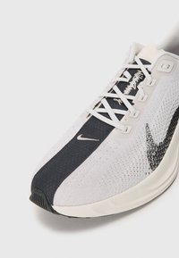 Chaussure de running Nike en blanc et noir avec une tige en tricot, des lacets blancs et un swoosh de marque sur le côté et sur la languette, sur fond blanc uni.