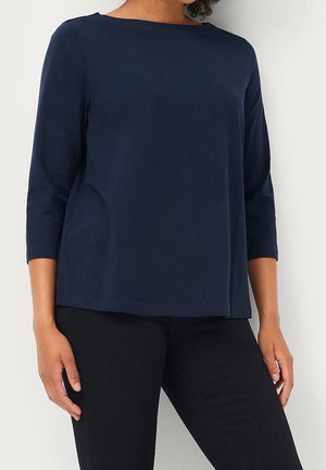 Personne portant un haut bleu marine uni à manches longues et un pantalon noir, debout devant un fond blanc, les mains détendues le long du corps.