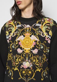 Black sweatshirt featuring a vibrant floral and baroque pattern in gold, pink, and blue with intricate details, including a sun motif and crown.
