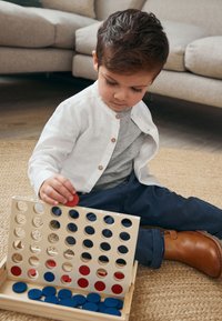 Gioco del quattro in fila in legno con pedine rotonde rosse e blu. Un bambino con una maglietta bianca e stivali marroni sta posizionando una pedina rossa nel gioco.