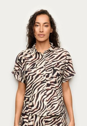 Donna con capelli scuri e ricci che indossa una camicia a maniche corte a righe zebrate con bottoni e pantaloni abbinati su uno sfondo chiaro e uniforme.