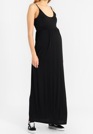 Gravid kvinde iført en ærmeløs sort maxikjole med tynde stropper og sort-hvide sneakers, stående mod en ensfarvet baggrund.