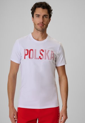Mężczyzna z kręconymi włosami, ubrany w białą koszulkę z czerwonym tekstem "POLSKA" i czerwonymi szortami, stoi na szarym tle.