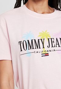 Camiseta de manga corta en rosa claro con "TOMMY JEANS" en texto negro en negrita, gráficos coloridos de palmeras y un acento tricolor en la parte inferior.
