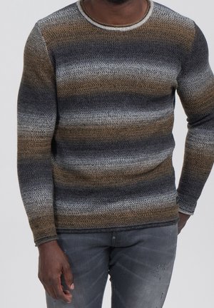 Homme portant un pull en tricot rayé marron, gris et noir, avec un jean en denim noir délavé, debout devant un fond uni.