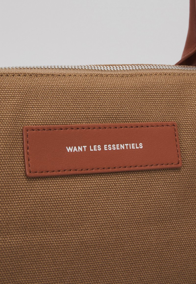 Braune Canvas-Tasche mit einem glatten Lederetikett, das in weißem Text "WANT LES ESSENTIELS" geprägt ist. Verfügt über einen silbernen Reißverschluss.