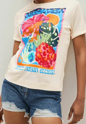 Personne portant un t-shirt beige avec un motif coloré de fruits tropicaux et de plantes et un texte bleu, associé à un short en jean bleu déchiré.