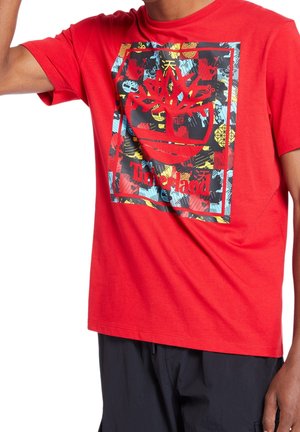 Red Timberland T-shirt with a colorful abstract square graphic and logo worn by a person in black pants against white background.
