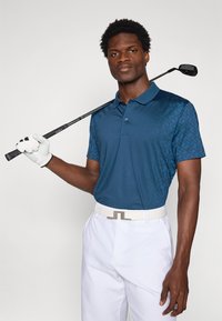 Navy blue golf polo with a textured pattern on the left side, paired with white trousers and a white belt, holding a black golf club.