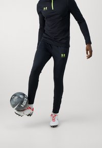 Svart atletisk långärmad tröja och åtsittande byxor med ljusgröna detaljer, kombinerat med vita och röda fotbollsdojor och en grå fotboll.