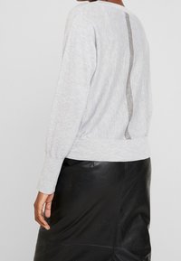 Maglione grigio chiaro a maniche lunghe, con una striscia verticale luccicante lungo la schiena e polsini a coste, abbinato a una gonna in pelle nera.