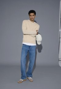 Beige hosszú ujjú kötött pulóver fehér ing fölött, világos kék farmernadrággal és szürke velúrbőr sportcipővel, krém színű sapkát tartva.