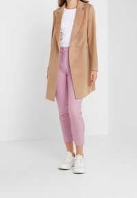 Lichtbruine dubbelgebreide jas, wit t-shirt eronder, roze geruite high-waisted broek en witte sneakers. Eenvoudig, strak ontwerp.