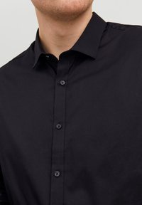 Camicia in cotone nera con colletto classico, caratterizzata da bottoni abbinati e una superficie liscia. Inquadratura ravvicinata sul colletto e sulla parte superiore del petto.