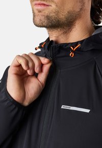 Schwarze Zip-Jacke mit verstellbarem Kapuzen, ausgestattet mit orangefarbenen Kordeln, einem kleinen weißen Logo auf der Brust und strukturiertem Stoff für zusätzliche Haltbarkeit.