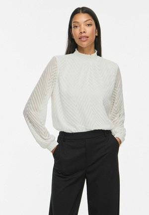 Femme portant un chemisier blanc texturé à manches longues avec col montant et un pantalon noir taille haute, debout les mains dans les poches.