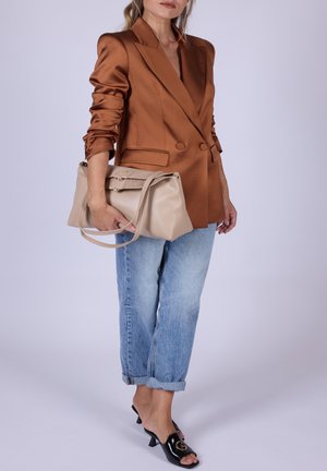 Donna con blazer in satin bronzo, jeans azzurri risvoltati, tacchi neri aperti in punta, che tiene una borsa in pelle beige con tracolla.